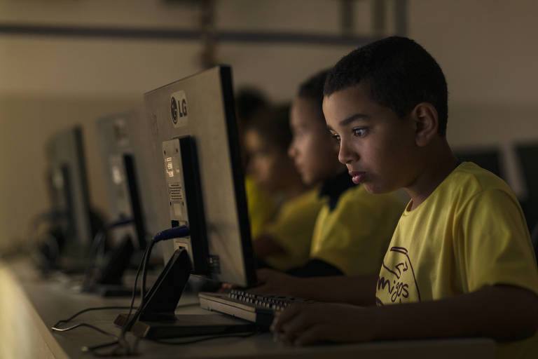 A imagem mostra um grupo de crianças sentadas em frente a computadores em uma sala com pouca iluminação. Um menino em destaque, usando uma camiseta amarela, está concentrado na tela do computador. Outros meninos podem ser vistos ao fundo, também utilizando os computadores. O ambiente parece ser uma sala de aula de informática.