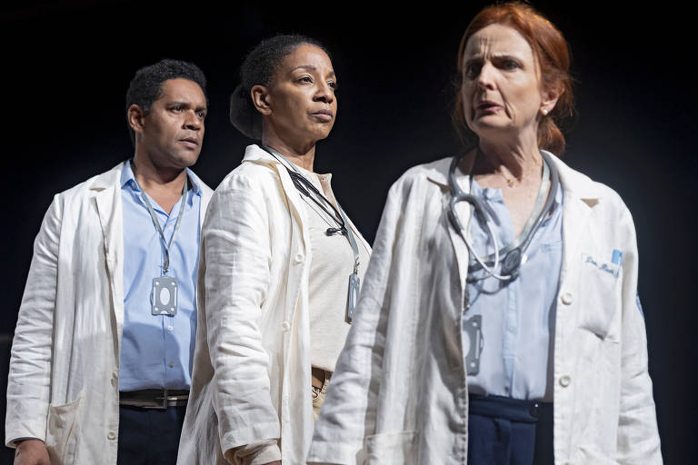 A imagem mostra três pessoas em um palco, vestindo jalecos brancos de médicos. O homem à esquerda tem cabelo cacheado e uma expressão séria. A mulher no centro, com cabelo preso, também tem uma expressão neutra. A mulher à direita, com cabelo ruivo, parece preocupada. Todos estão usando estetoscópios e crachás de identificação. O fundo é escuro, destacando os personagens.