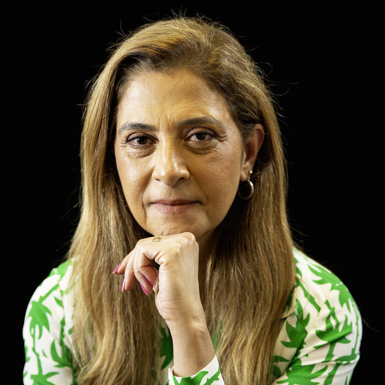 A imagem mostra uma mulher com cabelo longo e liso, usando uma blusa branca com estampas verdes. Ela está olhando diretamente para a câmera, com uma expressão séria, enquanto apoia o queixo com a mão. O fundo da imagem é preto, destacando a figura da mulher.