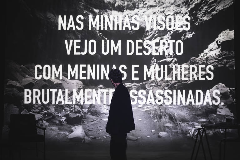 A imagem mostra uma pessoa de costas, em um ambiente escuro, diante de uma projeção que exibe um texto em letras grandes e brancas. O texto diz: 'NAS MINHAS VISÕES VEJO UM DESERTO COM MENINAS E MULHERES BRUTALMENTE ASSASSINADAS.' Ao fundo, há uma paisagem rochosa que complementa a atmosfera sombria da cena. No lado esquerdo, há uma cadeira e, à direita, uma mesa com outra cadeira.