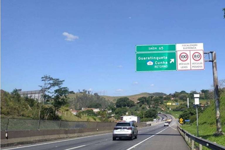 Imagem da estrada, com uma placa verde indicando a entrada de Guaratinguetá e um radar no acostamento à frente