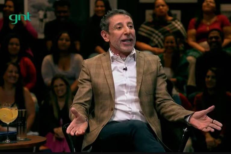 Um apresentador está sentado em uma cadeira, gesticulando com as mãos enquanto fala. Ele usa um paletó claro sobre uma camisa branca e calças escuras. À sua frente, há uma mesa com um copo de bebida. Ao fundo, uma plateia composta por várias pessoas, algumas sorrindo e outras prestando atenção. O ambiente parece ser um estúdio de gravação de um programa de televisão.