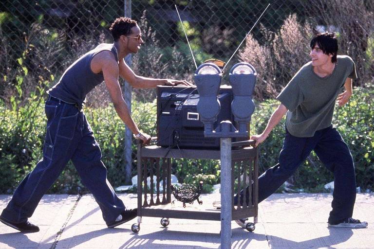 A imagem mostra dois jovens em uma rua. Um deles, à esquerda, está puxando um carrinho que contém um equipamento de som e dois parquímetros. O outro jovem, à direita, está empurrando o carrinho. Ambos estão vestidos de forma casual, com camisetas e calças largas. Ao fundo, há uma cerca e vegetação.