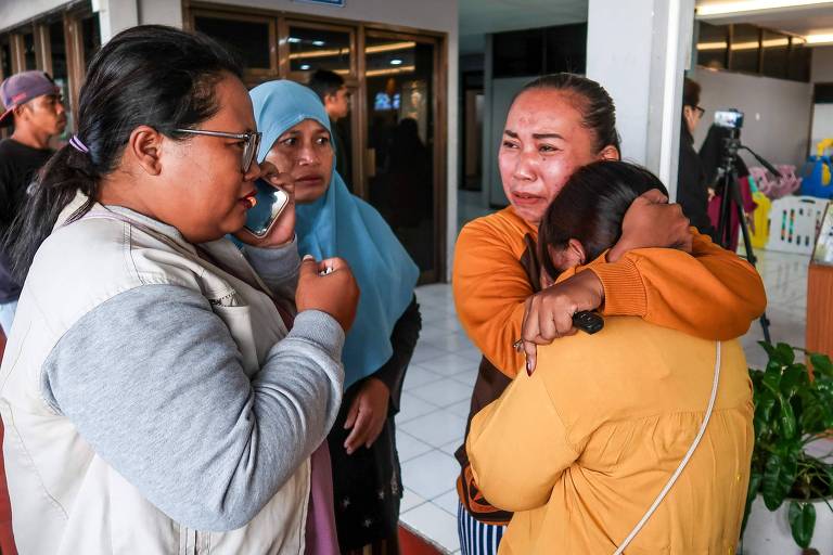 The image shows a group of four women indoors, apparently in a moment of mourning or sadness. One woman on the left is talking on the phone, while the other three are hugging each other and showing intense emotions, with tears visible on their faces. The room is well-lit and there are people in the background, but they are not clearly visible.