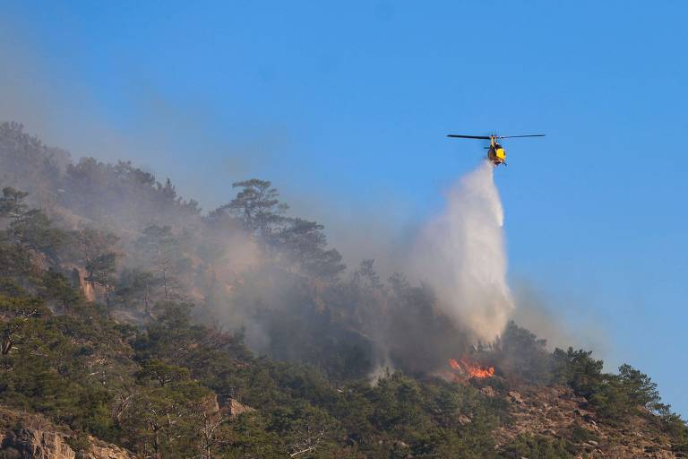 Um helicóptero amarelo está sobrevoando uma área montanhosa, despejando água sobre um incêndio florestal. A fumaça se eleva da vegetação em chamas, enquanto árvores verdes estão visíveis ao redor. O céu está claro e azul, contrastando com a cena de incêndio.