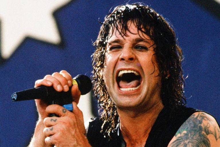 Um cantor está em um palco, segurando um microfone e cantando com intensidade. Ele tem cabelo longo e molhado, e está usando uma camiseta preta. O fundo é desfocado, mas parece ter elementos de um show, como luzes e uma bandeira ou pano com estrelas.