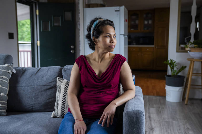 Uma mulher está sentada em um sofá cinza, olhando para o lado. Ela usa uma blusa vermelha e jeans. O ambiente ao fundo é uma sala de estar com uma porta aberta, uma geladeira branca e uma cozinha visível. Há plantas em vasos e almofadas decorativas no sofá.