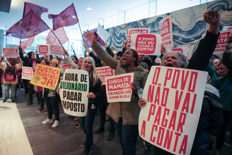 A imagem mostra um grupo de pessoas em um protesto, segurando cartazes e bandeiras. Os cartazes contêm mensagens como 'O POVO NÃO VAI PAGAR A CONTA', 'HORA DE BILIONÁRIO PAGAR IMPOSTO', e 'JUSTIÇA TRIBUTÁRIA JÁ!'. As pessoas estão visivelmente engajadas, algumas levantando os braços e outras gritando. O ambiente parece ser um espaço público, possivelmente um edifício governamental ou uma praça.