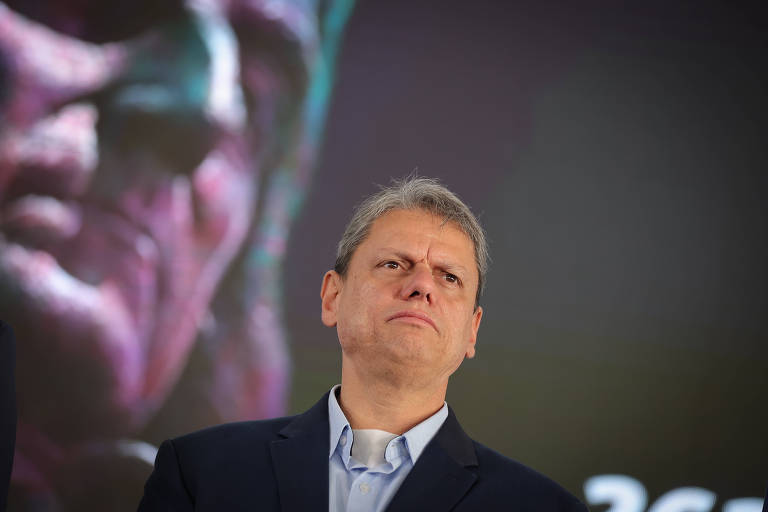 A man with short, gray hair, wearing a dark suit jacket and light-colored shirt, stands with a serious expression. In the background is an artistic image of a figure in shades of purple and green, which appears to be a sculpture or a painting. The lighting is soft, and the setting appears to be a formal event.