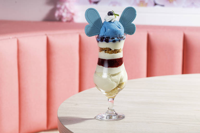 Uma taça de sobremesa com camadas de sorvete, creme e gelatina. O sorvete azul em cima é decorado com uma pequena folha e frutas escuras. A taça é transparente e está sobre uma mesa de madeira clara, com um fundo de sofá rosa.
