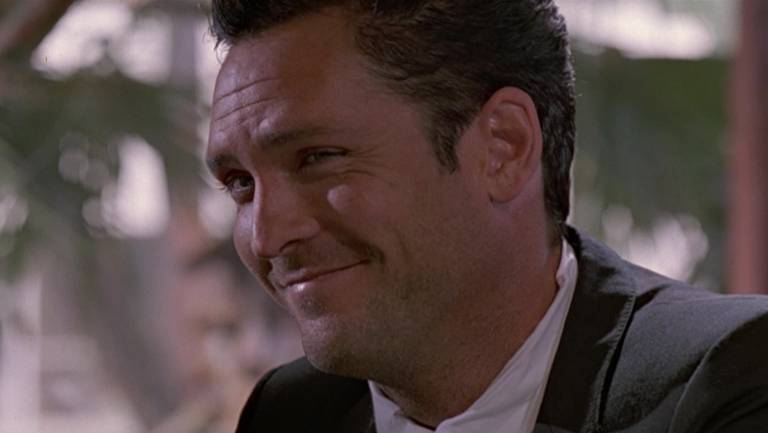 A imagem mostra um homem sorrindo, vestindo um terno escuro com uma camisa branca. Ele tem cabelo escuro e está em um ambiente que parece ser um restaurante ou bar, com um fundo desfocado que sugere uma atmosfera social. O homem parece estar em um momento descontraído e amigável.
