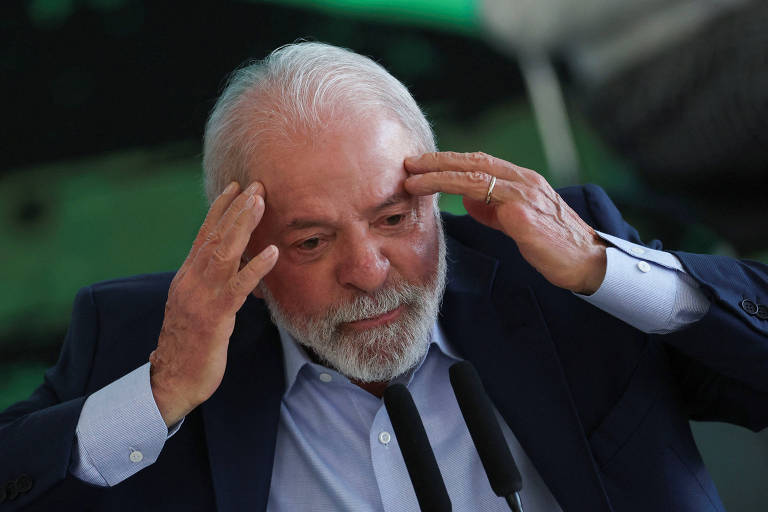 Um homem de cabelos grisalhos e barba, vestindo um paletó escuro e uma camisa clara, está com as mãos na cabeça, demonstrando uma expressão de preocupação ou estresse. Ele está em um ambiente que parece ser um evento público, com microfones à sua frente e um fundo desfocado que sugere uma plateia ou um cenário institucional.