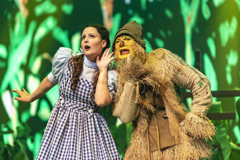 A imagem mostra dois personagens em uma cena de teatro. À esquerda, uma mulher com um vestido azul e branco, com cabelo preso em duas tranças, parece estar escutando atentamente. À direita, um personagem vestido como um espantalho, com uma máscara facial e roupas de palha, está inclinado em direção à mulher, como se estivesse sussurrando algo. O fundo é verde e apresenta um ambiente que remete a um cenário de floresta.