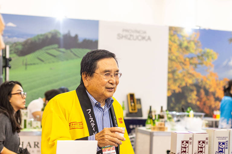 A imagem mostra um evento ou feira onde um homem vestido com um casaco amarelo e uma faixa preta está conversando com um grupo de pessoas. O fundo apresenta uma paisagem verde e montanhas, possivelmente representando a região de Shizuoka, no Japão. Há também produtos expostos em uma mesa, incluindo garrafas e embalagens. As pessoas ao redor parecem interessadas na conversa e nos produtos.