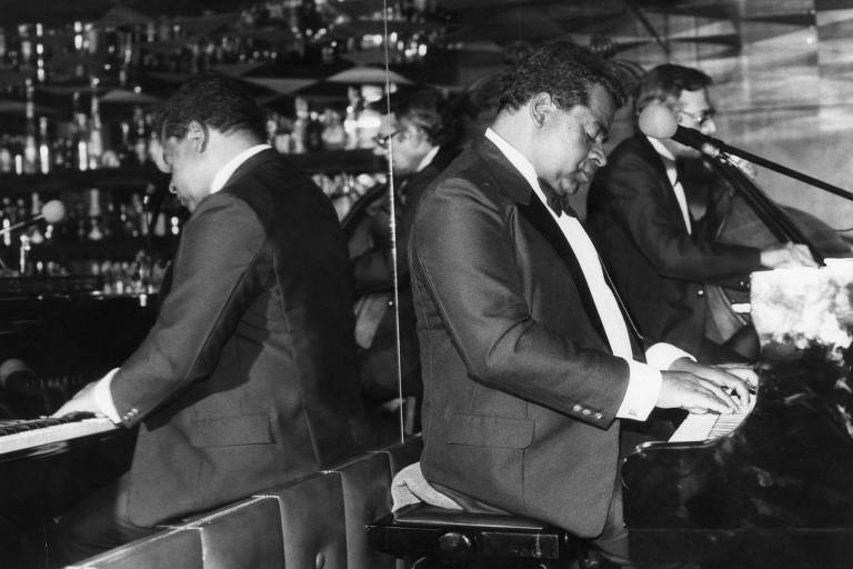 A imagem em preto e branco mostra um músico tocando piano em um ambiente de bar ou casa de shows. Ele está vestido com um terno escuro e uma camisa clara, concentrado em sua performance. Ao fundo, há um reflexo em um espelho e outros músicos visíveis, incluindo um que toca contrabaixo. O ambiente é iluminado com prateleiras de bebidas ao fundo.