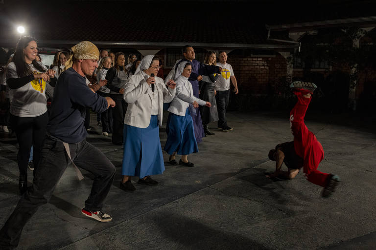A imagem mostra um grupo de pessoas participando de uma apresentação de dança em um ambiente externo à noite. Algumas pessoas estão vestidas com camisetas brancas e outras com roupas de freiras. Um dançarino em destaque está fazendo uma manobra de dança no chão, enquanto os outros observam e se divertem. O cenário é iluminado por luzes artificiais, criando um ambiente festivo.