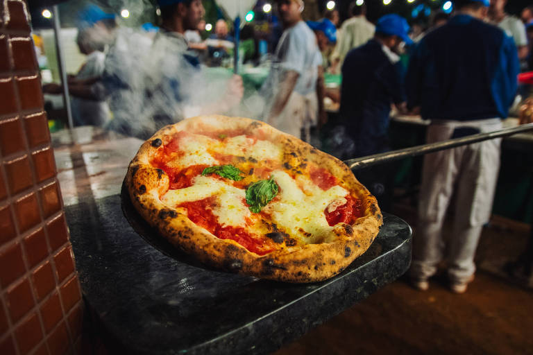 Uma pizza recém-assada, com queijo derretido e folhas de manjericão, está sendo retirada de um forno. A pizza é colocada em uma superfície escura e está soltando vapor. Ao fundo, várias pessoas estão em um ambiente de festa ou evento, algumas usando bonés azuis, e há mesas com comida.