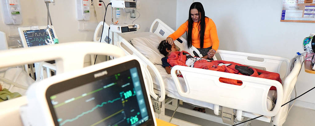 A imagem mostra um quarto de hospital com um paciente deitado em uma cama hospitalar. O paciente está coberto com um cobertor vermelho e parece estar recebendo cuidados. Ao lado do paciente, uma pessoa está sentada, possivelmente um familiar ou cuidador. Em primeiro plano, há um monitor de sinais vitais exibindo gráficos e números, indicando a condição do paciente. O ambiente é bem iluminado e organizado, com equipamentos médicos visíveis ao fundo.