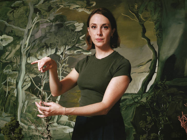 A imagem mostra uma mulher em pé, com cabelo castanho claro e liso, usando uma camiseta verde e uma saia escura. Ela está em um ambiente que parece ser uma pintura de fundo com árvores e um cenário natural. A mulher está gesticulando com a mão direita, segurando algo com a mão esquerda, enquanto olha para a câmera.