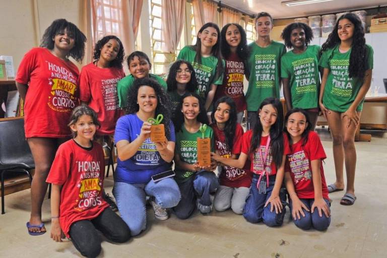 A imagem mostra um grupo de jovens e uma mulher adulta em um ambiente interno. Eles estão sorrindo e posando para a foto. As jovens estão vestidas com camisetas coloridas, principalmente em vermelho e verde, com frases impressas. A mulher adulta, que está agachada no centro, segura dois troféus ou prêmios. O ambiente parece ser uma sala de aula ou um espaço comunitário, com janelas grandes ao fundo e prateleiras com livros.