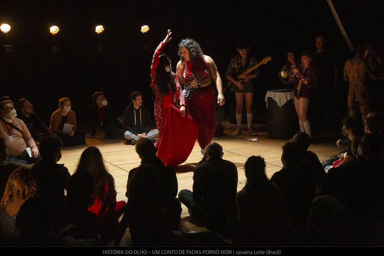 A imagem mostra uma apresentação de dança em um palco iluminado. Duas dançarinas vestidas com trajes vermelhos estão no centro do palco, dançando uma para a outra. O público, composto por várias pessoas sentadas em círculo ao redor do palco, observa atentamente. Ao fundo, há músicos com instrumentos, e uma mesa coberta com um pano branco. A atmosfera é de um evento cultural, com foco na performance das dançarinas.