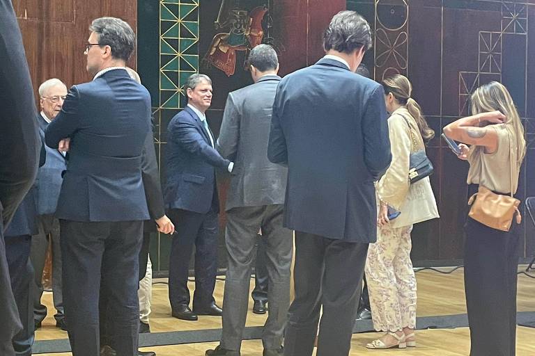 A imagem mostra um grupo de pessoas em um ambiente interno, possivelmente em um evento social ou reunião. A maioria das pessoas está vestida formalmente, com trajes escuros. Algumas estão conversando entre si, enquanto outras parecem estar se preparando para se mover. O fundo apresenta uma parede decorada com elementos gráficos e cores variadas, sugerindo um ambiente elegante. O piso é de madeira clara.