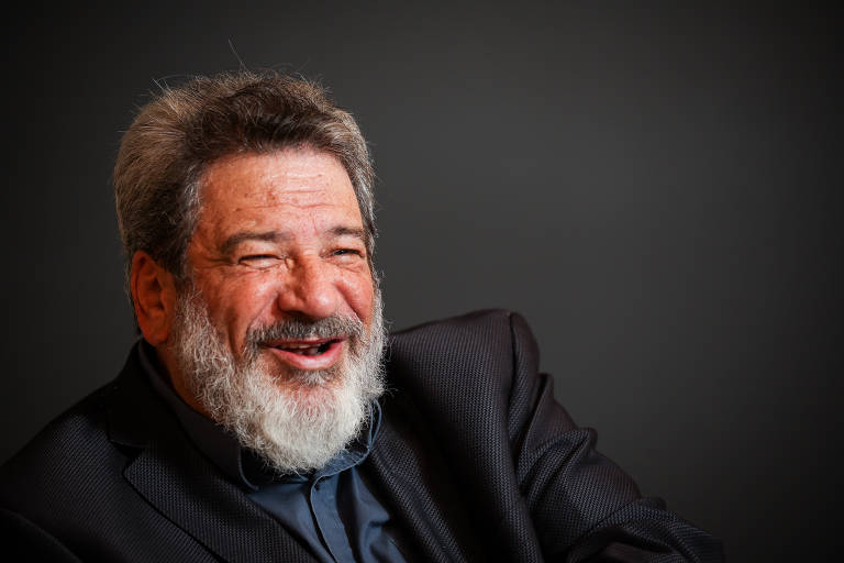 Um homem com cabelo grisalho e barba, vestindo um terno escuro, está sorrindo. O fundo é de cor escura, destacando a expressão amigável do homem.