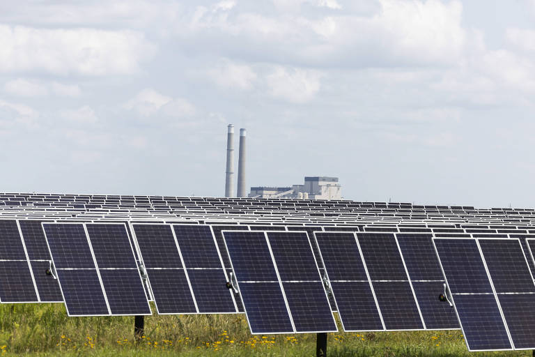 A imagem mostra uma grande instalação de painéis solares dispostos em fileiras, com um céu claro e algumas nuvens ao fundo. Ao fundo, é possível ver uma estrutura industrial com chaminés, sugerindo a presença de uma usina. A vegetação ao redor é baixa e há algumas flores amarelas visíveis.