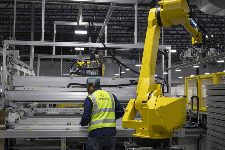 A imagem mostra um ambiente industrial com um trabalhador usando um colete refletivo amarelo e um boné, operando próximo a uma máquina. À direita, há um robô amarelo com um braço articulado, que parece estar em operação. O fundo é composto por estruturas metálicas e iluminação artificial, característico de uma fábrica moderna.