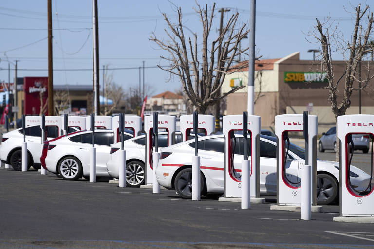 A imagem mostra uma fila de carros Tesla estacionados em uma estação de carregamento. Os veículos são predominantemente brancos e estão alinhados ao lado de carregadores Tesla, que são estruturas brancas com detalhes vermelhos. Ao fundo, é possível ver alguns estabelecimentos comerciais e árvores sem folhas, indicando que a foto foi tirada em um dia claro e ensolarado.