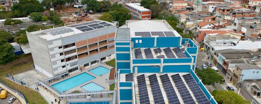 A imagem mostra uma escola moderna com telhados equipados com painéis solares. A estrutura é composta por vários andares e possui uma quadra de esportes em primeiro plano, com um campo de basquete. Ao fundo, é possível ver uma área urbana com várias casas e edifícios. O céu está nublado, e a imagem foi capturada de uma perspectiva aérea.