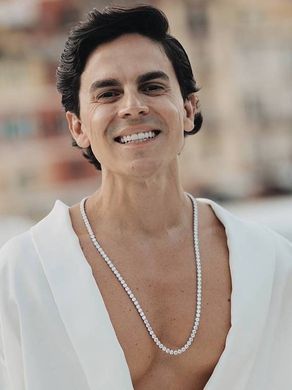 Um homem sorridente está posando em um ambiente ao ar livre, com um fundo de casas coloridas à beira-mar. Ele usa um robe branco aberto, revelando uma camiseta ou parte do torso, e está adornado com um colar de pérolas. O homem tem cabelo escuro e liso, e sua expressão é alegre. O cenário sugere um clima de verão e descontração.