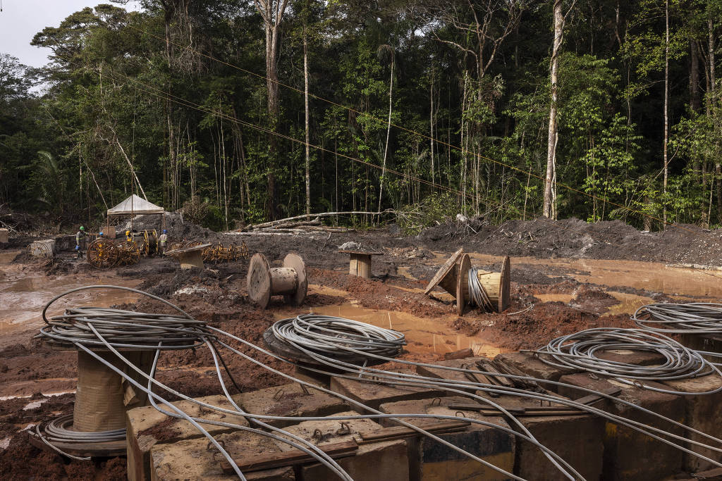 A imagem mostra uma área de extração em uma floresta, com várias bobinas de cabos e fios no primeiro plano. O solo está coberto de lama e há uma estrutura de lona ao fundo, onde trabalhadores estão visíveis. A vegetação ao redor é densa, com árvores altas e verdes, indicando um ambiente de floresta tropical.