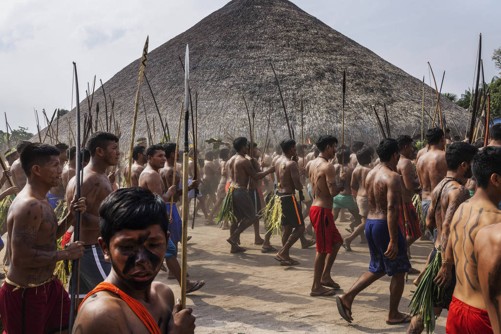 A imagem mostra um grupo de homens indígenas, sem camisa, participando de uma cerimônia. Eles estão armados com lanças e segurando folhas. Ao fundo, há uma grande estrutura de telhado de palha, típica de habitações indígenas. O ambiente é ao ar livre, com um céu claro e algumas nuvens.