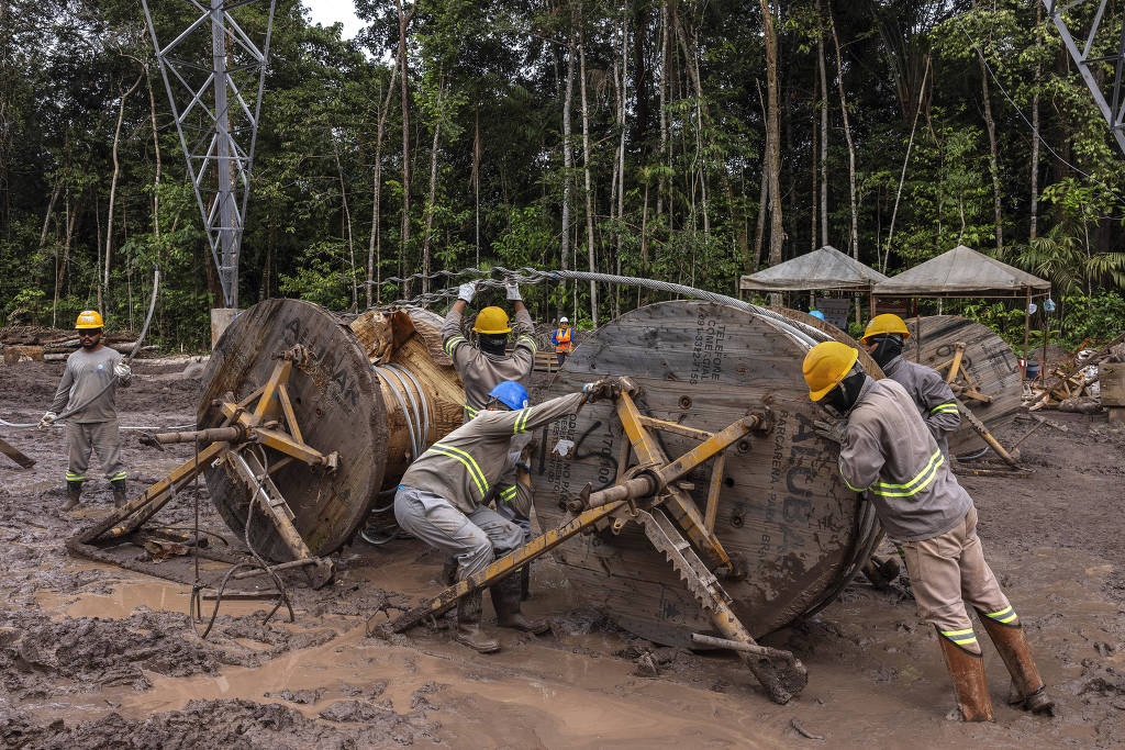 A imagem mostra um grupo de trabalhadores em um local de construção, cercados por árvores e vegetação densa. Eles estão usando capacetes de segurança e roupas de trabalho, enquanto tentam mover grandes bobinas de cabo. O solo está lamacento, indicando que o trabalho está sendo realizado em condições desafiadoras. Ao fundo, há torres de energia e estruturas cobertas.