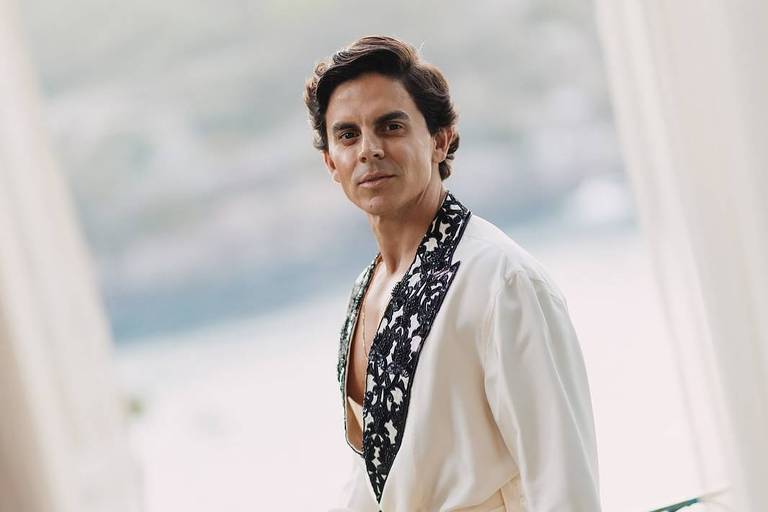 Um homem está posando em uma varanda, vestindo uma camisa branca com um detalhe escuro e um lenço estampado. Ele tem cabelo escuro e está olhando para a câmera com uma expressão confiante. Ao fundo, pode-se ver uma paisagem desfocada, possivelmente um corpo d'água e vegetação. A luz é suave, sugerindo um ambiente tranquilo e sofisticado.