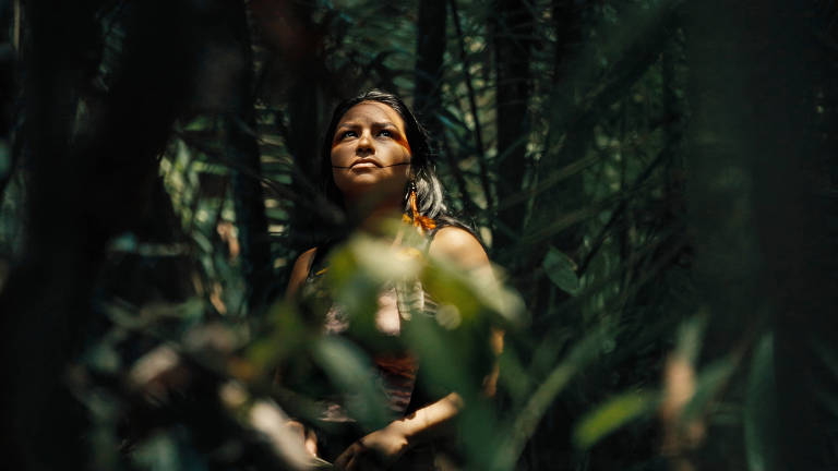 A imagem mostra uma mulher em um ambiente de floresta densa, cercada por folhas verdes. Ela está olhando para cima, com uma expressão contemplativa. A luz natural ilumina seu rosto, destacando seus traços. A vegetação ao redor é rica e variada, criando um fundo escuro que contrasta com a figura da mulher.