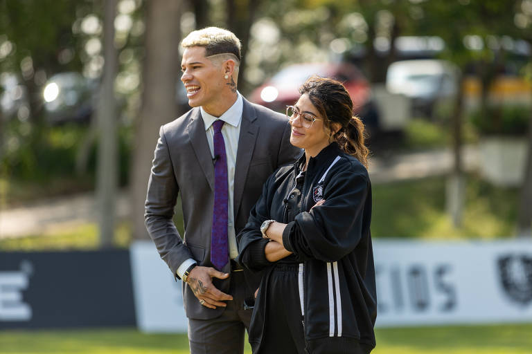 Um homem e uma mulher estão sorrindo e conversando em um evento ao ar livre. O homem está vestido com um terno cinza e uma gravata roxa, enquanto a mulher usa um casaco preto e óculos de sol. Ao fundo, há árvores e veículos estacionados.