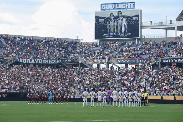 A imagem mostra um estádio de futebol lotado, com torcedores nas arquibancadas. No campo, duas equipes estão alinhadas em uma cerimônia, possivelmente em homenagem. Um telão ao fundo exibe imagens de dois jogadores. O céu está parcialmente nublado e há uma grande faixa com o nome 'BRAVO1952' visível nas arquibancadas.