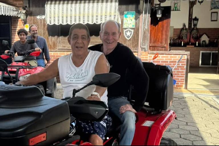Na imagem, duas pessoas estão sentadas em um quadriciclo vermelho. A pessoa à frente, com uma camiseta branca e calças curtas, está sorrindo. A pessoa atrás, com um suéter preto e calças jeans, também está sorrindo. Ao fundo, há outras pessoas e uma construção com detalhes em madeira. O ambiente parece ser ao ar livre, com um céu claro.
