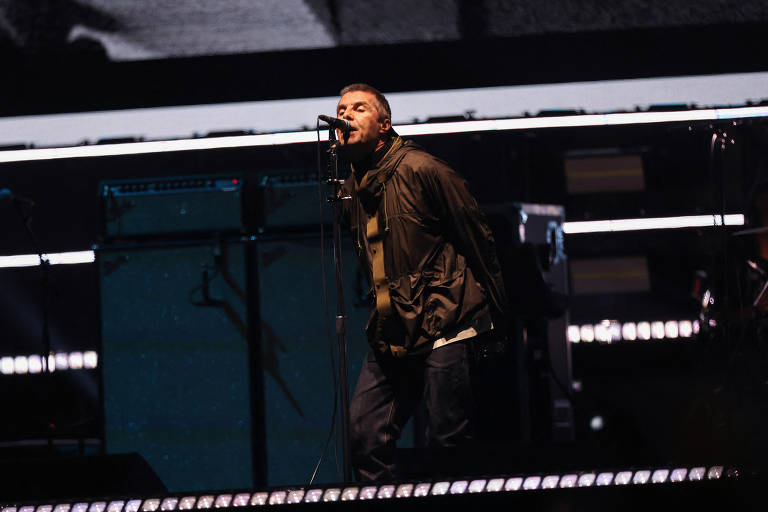 Um cantor está se apresentando em um palco, segurando um microfone e cantando com intensidade. Ele está vestido com uma jaqueta escura e calças jeans. Ao fundo, há amplificadores e luzes de palco. A atmosfera é de um show ao vivo, com foco na performance do artista.