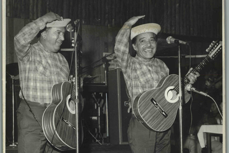 A imagem mostra dois homens em um palco, ambos usando camisas xadrez e chapéus. O homem à esquerda está segurando um violão e sorrindo, enquanto o homem à direita também sorri e levanta a mão em um gesto de saudação. Ambos estão em frente a microfones, com um fundo que sugere um ambiente de apresentação musical. O piso é de azulejos e há mesas ao fundo, indicando um local de show.