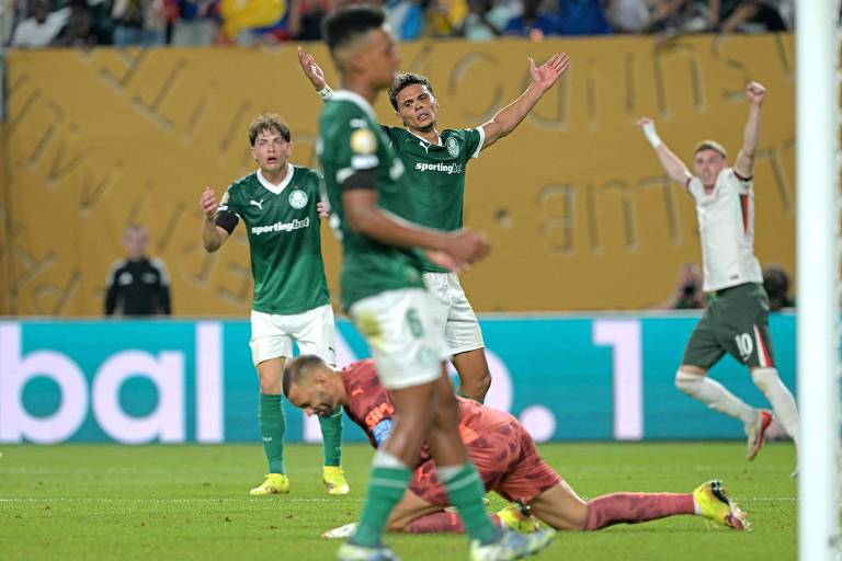 A imagem mostra um momento de um jogo de futebol. Jogadores estão em campo, alguns com expressões de frustração e outros comemorando. Um jogador está caído no chão, enquanto outros estão em pé, com um deles levantando os braços em sinal de reclamação. O fundo apresenta uma tela com informações do jogo e uma torcida ao fundo.