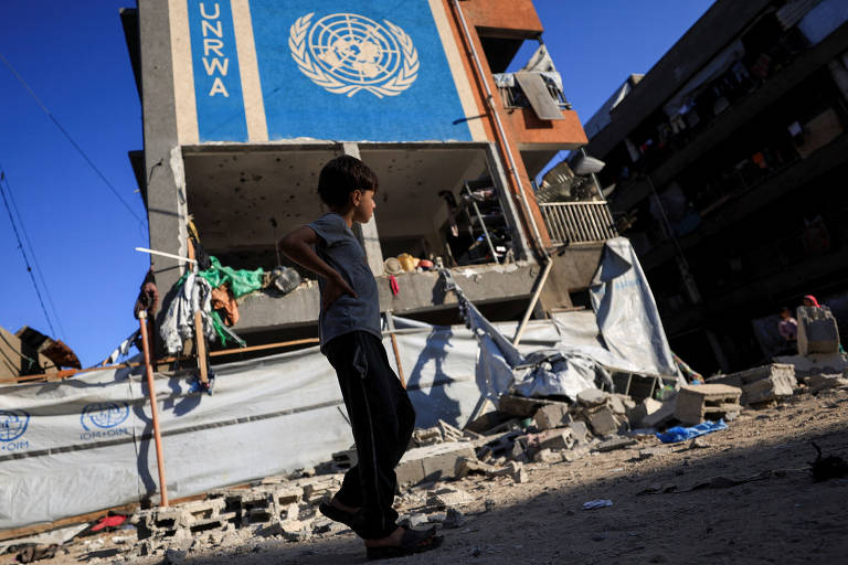 A imagem mostra uma criança de costas, vestindo uma camiseta cinza e calças escuras, caminhando em frente a um edifício da UNRWA (Agência da ONU de Assistência a Refugiados da Palestina) que apresenta danos visíveis. O prédio tem o logotipo da ONU em destaque na parte superior. Ao fundo, há escombros e uma estrutura de construção, com roupas penduradas em varais. O céu está claro e azul, indicando um dia ensolarado.