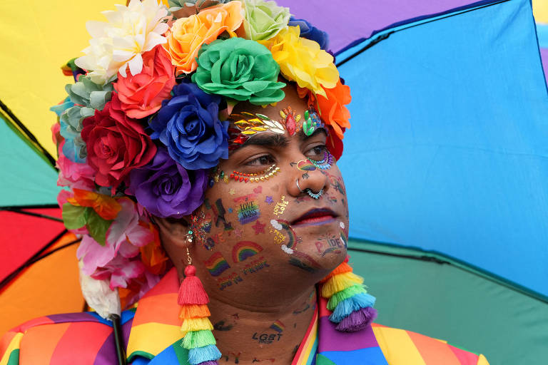 A imagem mostra uma pessoa com um rosto decorado com várias tatuagens coloridas e brilhantes, incluindo símbolos do arco-íris e a palavra 'LGBT'. A pessoa usa um chapéu ou tiara com flores coloridas e uma roupa com estampas em cores do arco-íris. Ao fundo, há um guarda-chuva também colorido em tons vibrantes. A expressão da pessoa é séria, e o ambiente sugere um evento de celebração da diversidade.