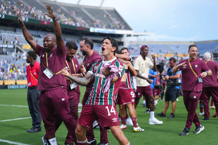 A imagem mostra uma cena de celebração de uma equipe de futebol após uma vitória. Vários jogadores e membros da comissão técnica estão em um campo, alguns levantando os braços e expressando alegria. Um jogador em destaque, usando a camisa número 14, está no centro da imagem, gritando e fazendo gestos de comemoração. O fundo mostra um estádio com torcedores.