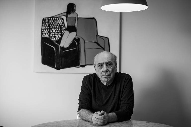 Um homem idoso está sentado à mesa, com as mãos cruzadas. Ele tem cabelo grisalho e uma expressão séria. Ao fundo, há uma pintura em preto e branco de uma mulher sentada em um sofá. A iluminação é suave, com uma lâmpada pendurada acima da mesa.