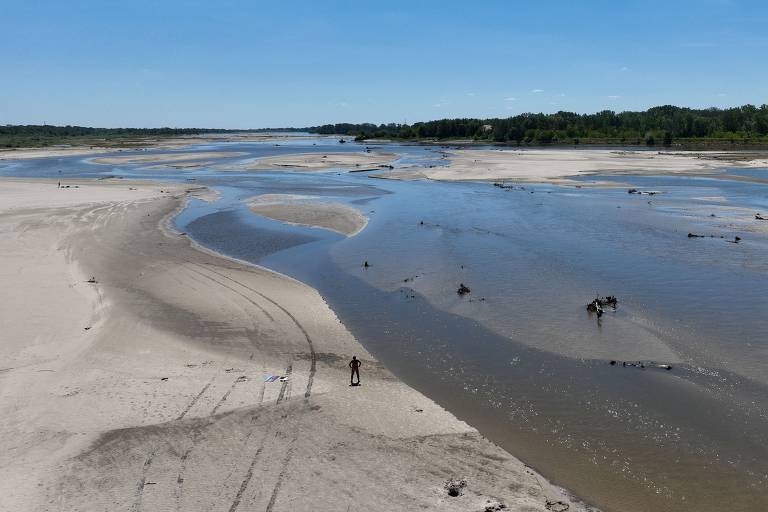 A imagem mostra uma ampla vista de um rio com áreas de areia exposta. O rio serpenteia pelo meio da paisagem, com águas calmas e algumas árvores ao fundo. Há uma pessoa em pé na areia, perto da margem do rio. O céu está claro e ensolarado, com poucas nuvens visíveis.