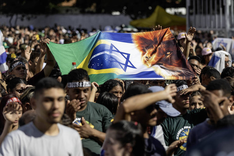 A imagem mostra uma grande multidão em um evento ao ar livre. Algumas pessoas estão levantando uma bandeira que combina elementos da bandeira do Brasil com a imagem de um leão. O público parece animado e envolvido, com várias pessoas visíveis em primeiro plano, algumas com expressões de entusiasmo. Ao fundo, há uma grande quantidade de pessoas, sugerindo um evento significativo.