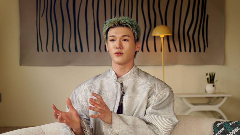 Um jovem com cabelo verde e roupas prateadas está sentado em um sofá, gesticulando enquanto fala. Ao fundo, há uma parede decorada com linhas verticais pretas e um abajur amarelo. O ambiente é moderno e minimalista, com uma mesa branca e uma planta em um vaso.